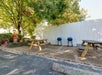 Outdoor patio area with two wooden picnic tables, two blue grills, a white fence, trees providing shade, and a scarecrow decoration near a pumpkin.