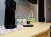 A coffee maker, two floral mugs, a TV remote, glass cups, and a basket with snacks sit on a yellow counter.