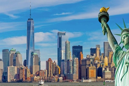 The Statue of Liberty stands with New York City skyscrapers in the background under a blue sky.