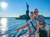 A couple poses by the waterfront with the Statue of Liberty in the background on a sunny day.
