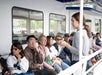 A woman stands and speaks to a group of seated people on a boat.