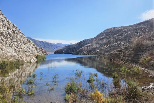 A serene lake surrounded by rocky hills under a clear blue sky, with sparse vegetation in the foreground.
