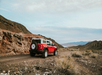 A red SUV with a white top drives on a dirt road through a rocky desert landscape under a cloudy sky.