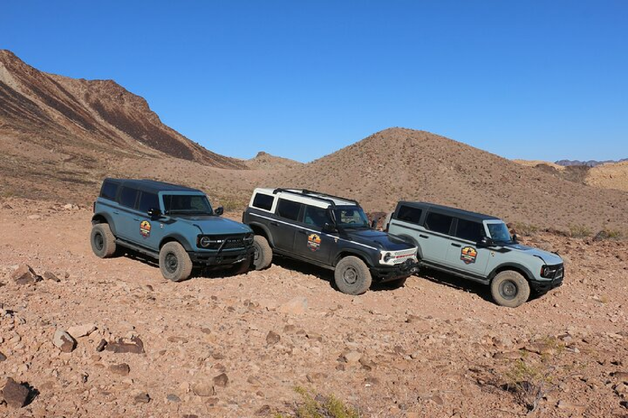 Three off-road vehicles parked on a rocky desert landscape with hills and a clear blue sky in the background.