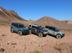 Three off-road vehicles parked on a rocky desert landscape with hills and a clear blue sky in the background.