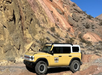 A yellow off-road vehicle with a white roof is parked on rocky terrain with steep, multicolored cliffs in the background.