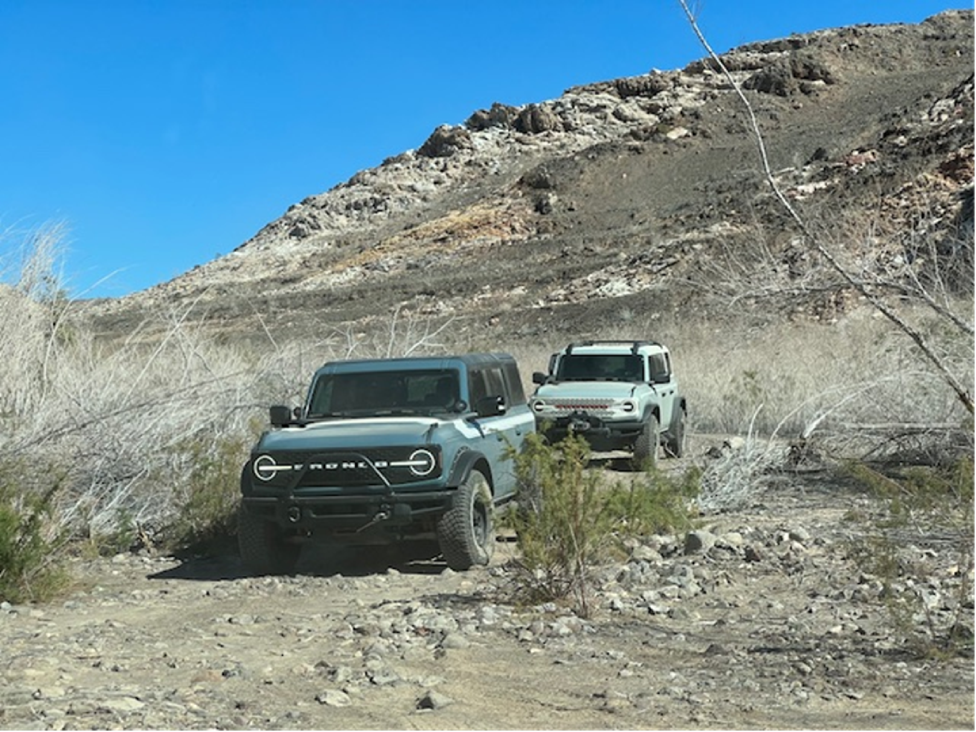 Two off-road vehicles drive on a rocky dirt trail surrounded by arid, mountainous terrain under a clear blue sky.