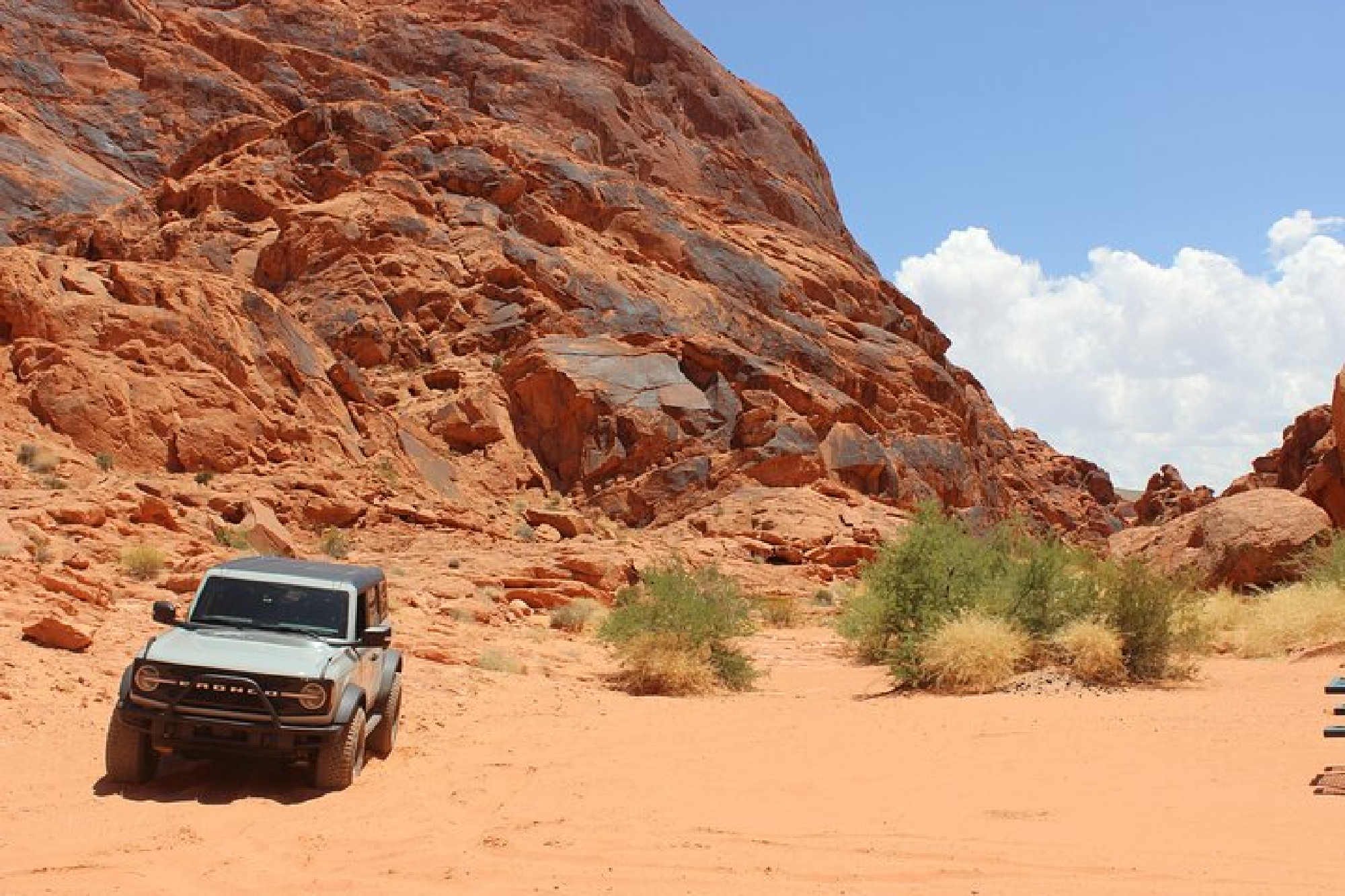 A vehicle navigates a sandy trail with red rock formations in a desert landscape under a clear blue sky.