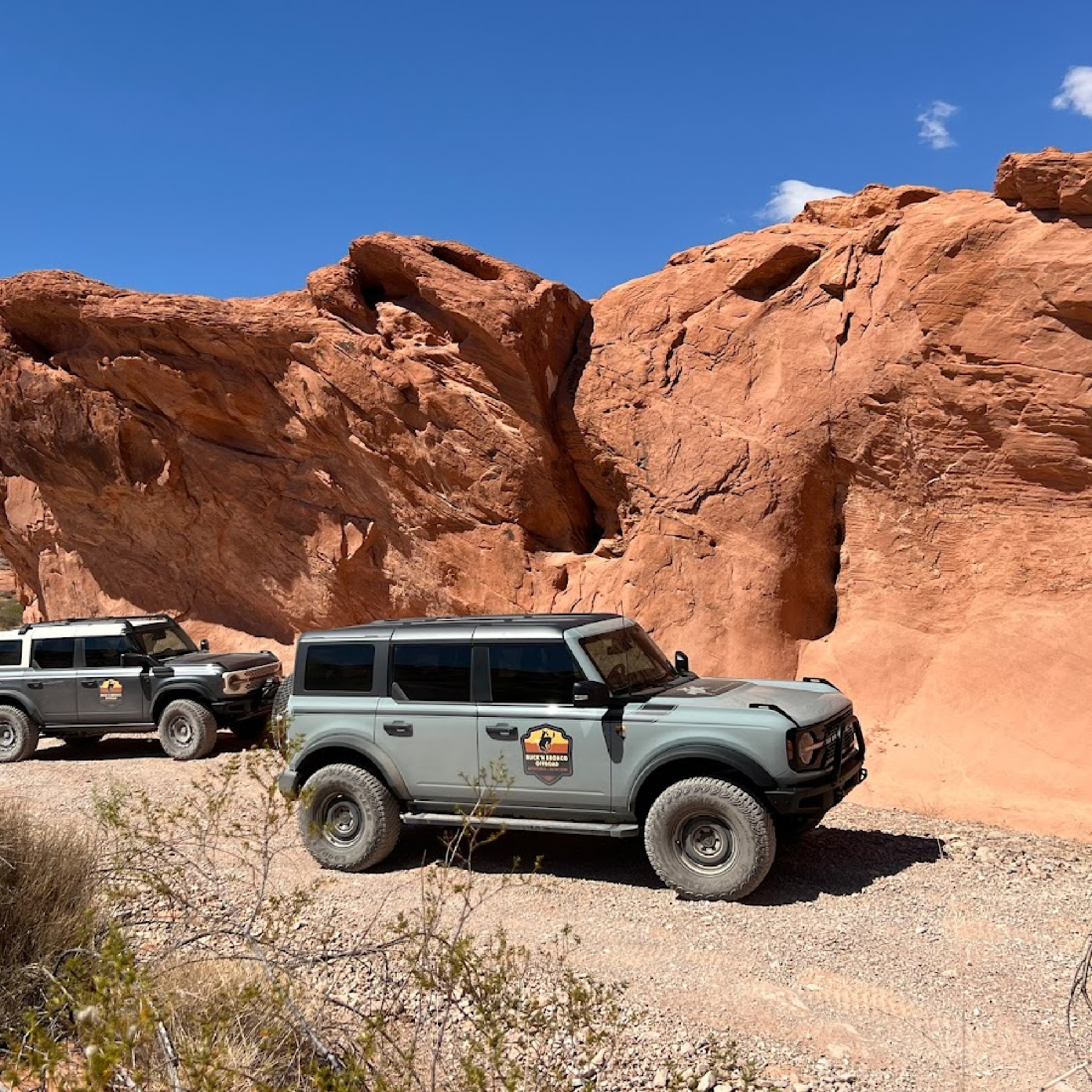 Two off-road vehicles parked on a gravel path in front of large red rock formations under a clear blue sky.