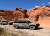 Two off-road vehicles parked on a gravel path in front of large red rock formations under a clear blue sky.