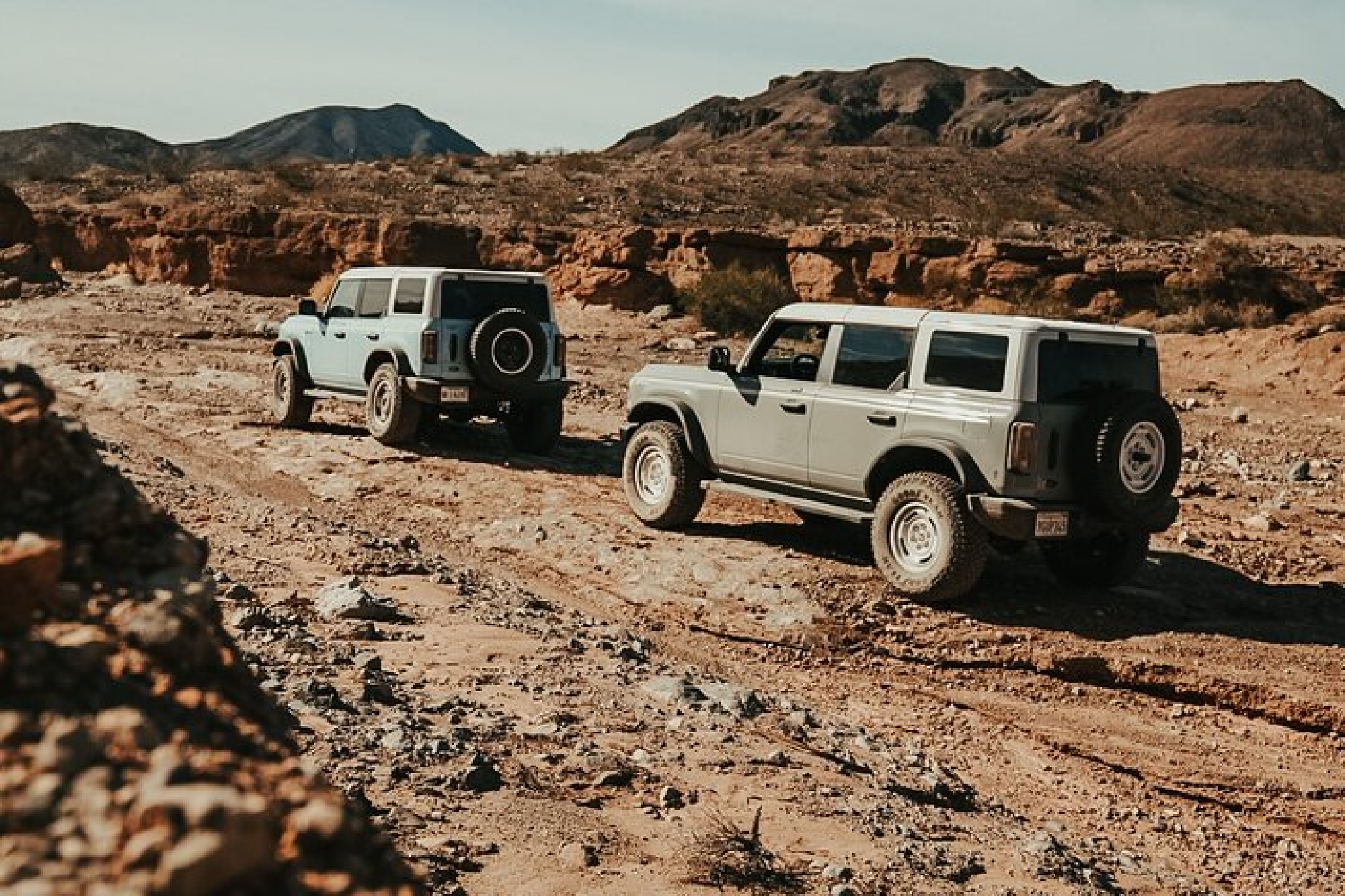 Two off-road vehicles drive through a rocky desert landscape with mountain views under a clear sky.