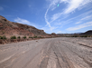 A dirt road stretches through a desert landscape surrounded by rocky hills under a blue sky with wispy clouds.