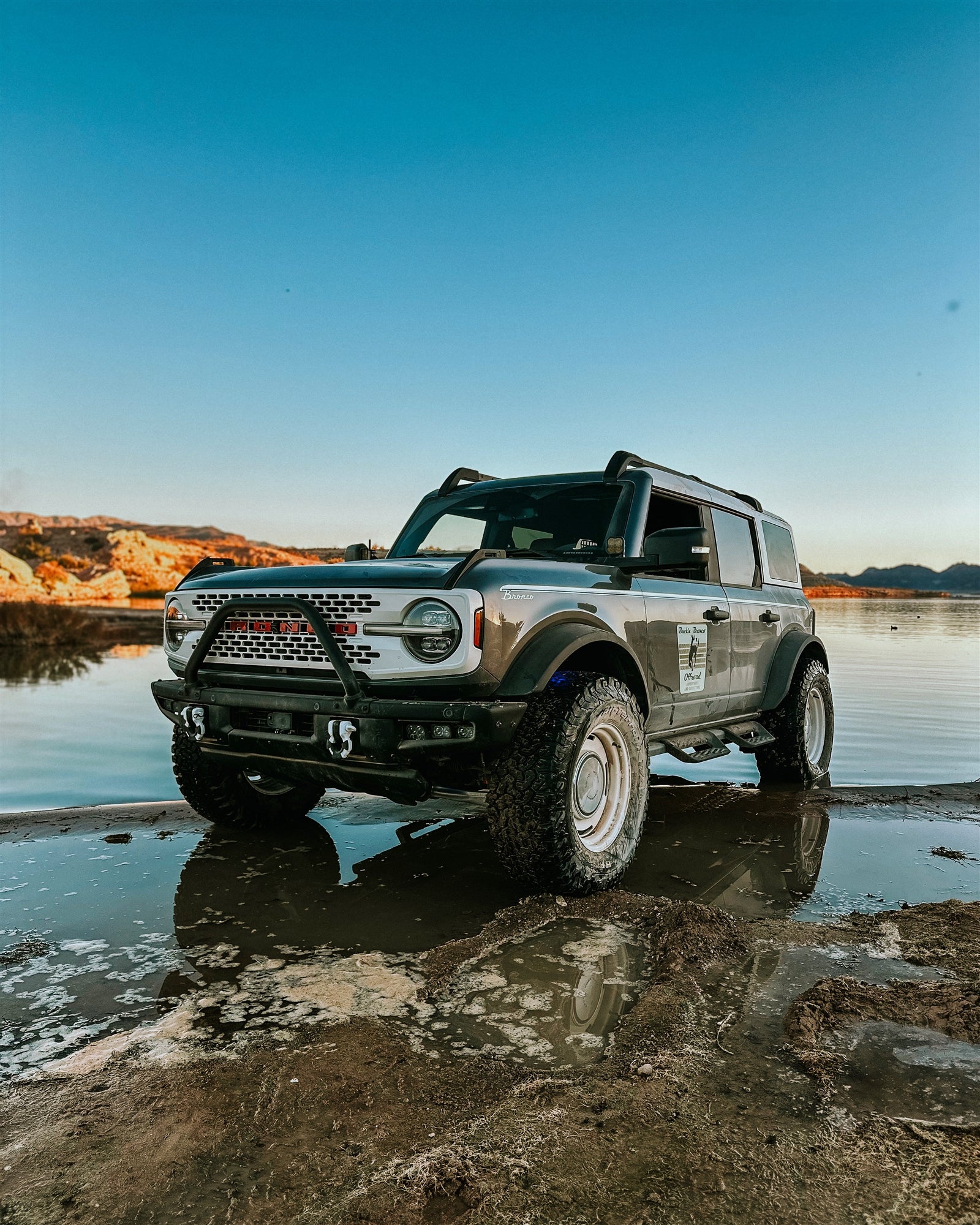 A gray off-road vehicle parked on muddy terrain near a body of water with a clear blue sky in the background.