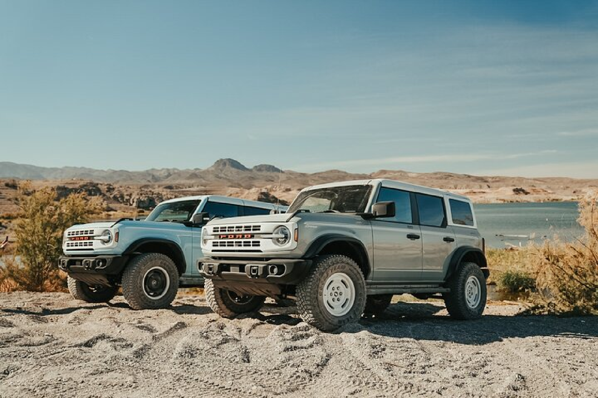 Two off-road vehicles parked on a sandy terrain with mountains and a body of water in the background under a clear blue sky.