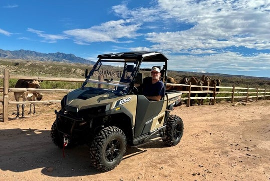 A person sits in an off-road vehicle on a dirt path near a fenced area with horses under a partly cloudy sky.