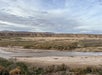 Desert landscape with a shallow river in the foreground, sparse vegetation, and distant hills under a cloudy sky.