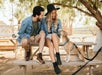 A man and woman sitting on a picnic table smile at a small dog. They are outdoors, wearing casual clothes and a hat.