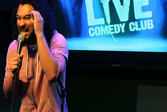 A comedian performs on stage holding a microphone, with a "Live Comedy Club" sign displayed in the background.