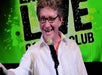 A man wearing glasses and a white shirt stands in front of a microphone, smiling, with a green and black "LIVE COMEDY CLUB" sign in the background.
