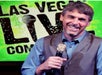 A man in a blue shirt and gray vest holding a microphone and performing at the Las Vegas Live Comedy Club with a green backdrop behind him.