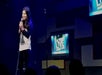 A woman stands on stage holding a microphone, performing stand-up comedy. Two screens behind her display the text "LIVE" in blue and white.