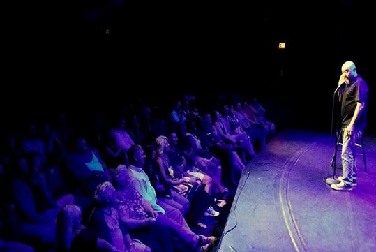 A man in a black shirt and jeans performing on stage speaking to a theatre full of people at the Las Vegas Live Comedy Club.
