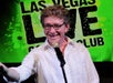 A man in a white button up shirt and glasses performing on stage at the Las Vegas Live Comedy Club with a lime green backdrop behind him.