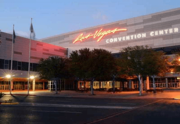 The front exterior of the Las Vegas Marriot with trees and flag poles in front and a glowing sign on the upper lever of it at dusk.