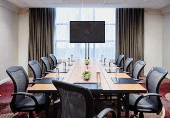 A modern conference room with a long wooden table with black office chairs around with a TV at the far end with a window behind it.