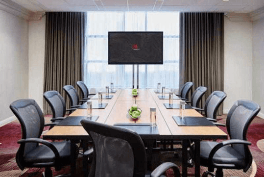 A modern conference room with a long wooden table with black office chairs around with a TV at the far end with a window behind it.