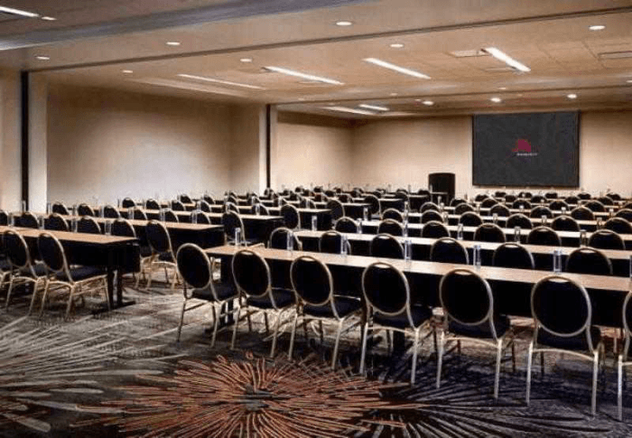 A conference room with rows of tables with black chairs lined up in front of a large projector screen at the front of the room.
