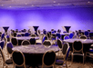 A banquet room with round black tables with black and silver chairs around them and purple lighting illuminating the room.