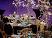 A banquet room with black tables set with an elaborate center piece and glassware with a purple wall behind them.
