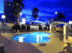 A small light blue outdoor swimming pool with lounge chairs around it at night with palm trees and a deep blue sky in the background.