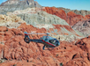 A gray helicopter with red accents flies over rugged red, white, and gray rock formations under a clear sky.