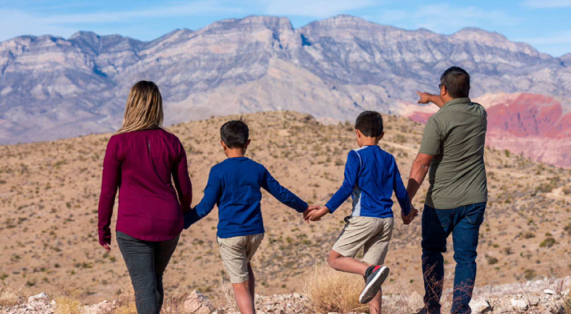 Four people, two adults and two children, stand on rocky terrain holding hands and looking towards distant mountains; one adult points ahead.