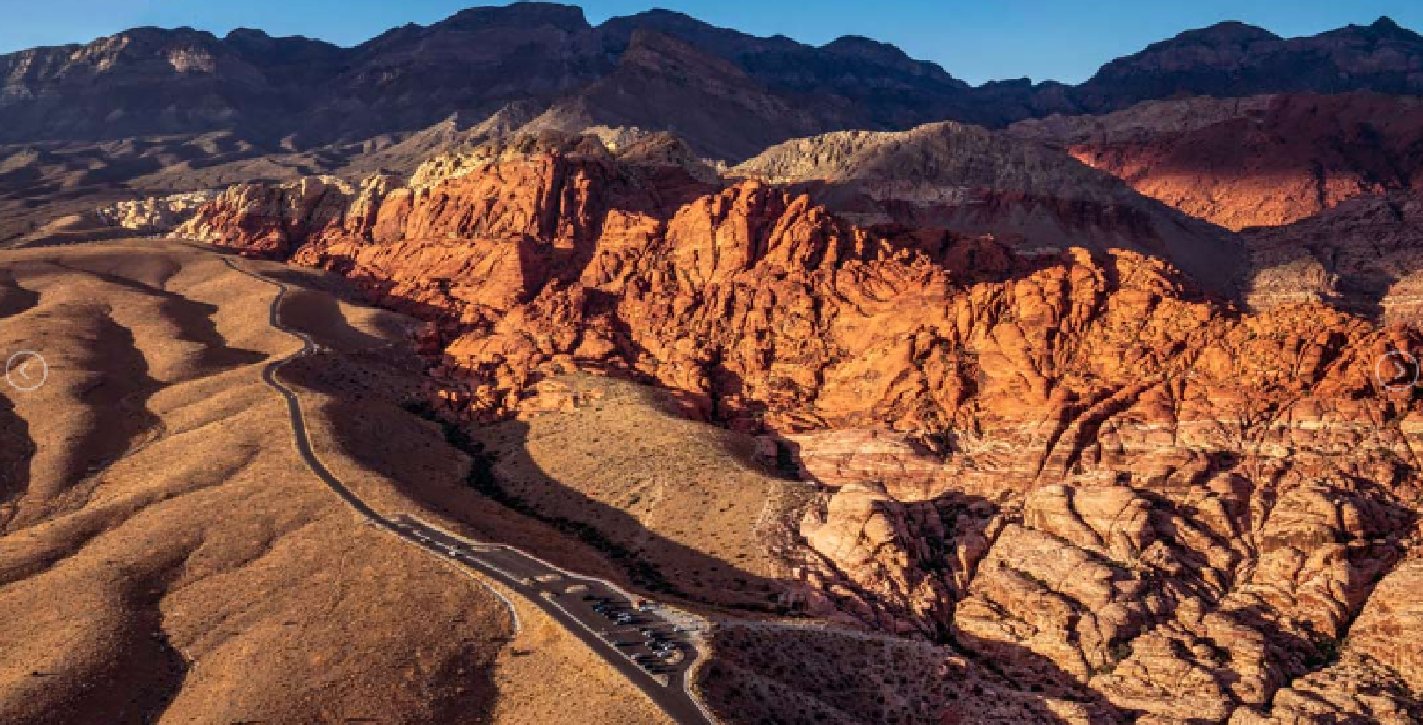 A winding road runs alongside rugged, red rock formations and mountains under a clear sky.
