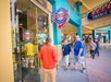 People walk past the entrance of Ultimate Sports Cards & Memorabilia store in an outdoor shopping center. Signs and business hours are visible on the glass door.