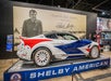 A Shelby American sports car with a patriotic paint design is displayed indoors on a platform in front of a large mural of Carroll Shelby.