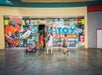 Three people walk past the entrance of a toy store with bright, colorful signage and various cartoon characters displayed on the window.