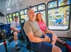 Three adults sit on a bus, smiling and looking out the window at a sunny street with palm trees and parked cars visible outside.