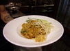 A plate of Pad Thai with noodles, bean sprouts, crushed peanuts, lime wedge, and green onion garnish, served on a white plate. A person with a compass tattoo holds the dish.