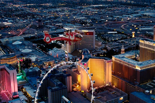 A red helicopter flies over the illuminated Las Vegas Strip at dusk, with the High Roller Ferris wheel and large casino hotels visible below.