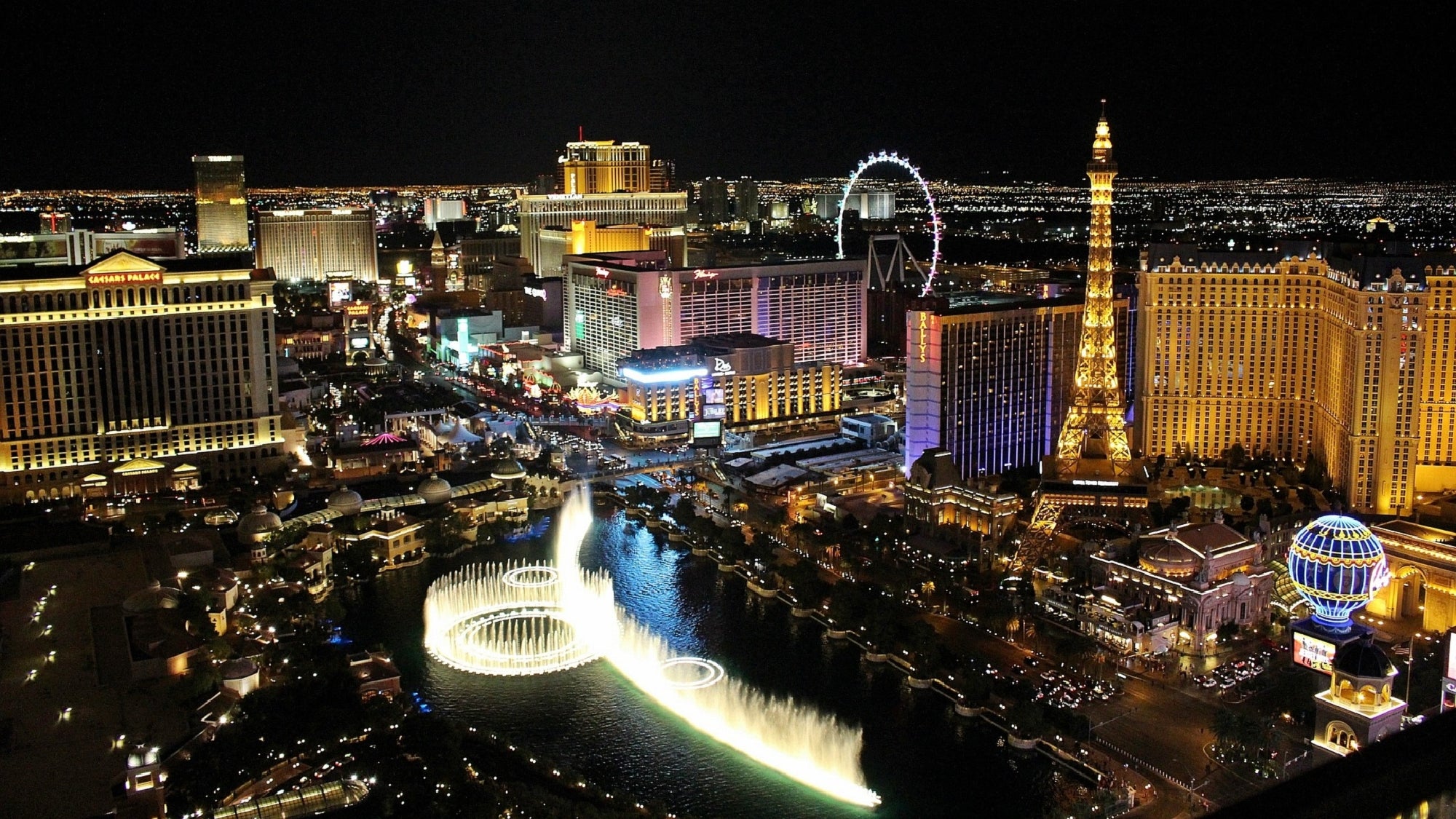 Aerial night view of the Las Vegas Strip, featuring brightly lit hotels, casinos, the Eiffel Tower replica, and the Bellagio fountain show in the foreground.