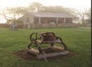 An old metal bell mounted on a wooden platform in a grassy yard, with a rustic wooden house and a picket fence in the background.