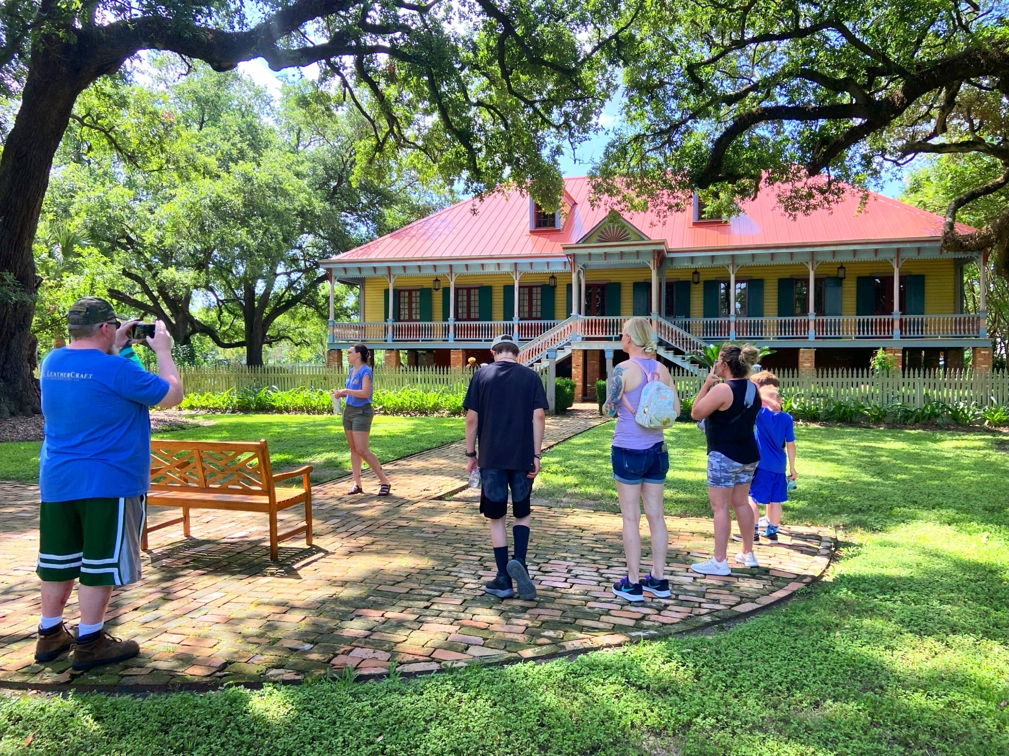 A group of people stands on a brick path in front of a large, historic house with a red roof and green shutters, surrounded by trees and greenery.