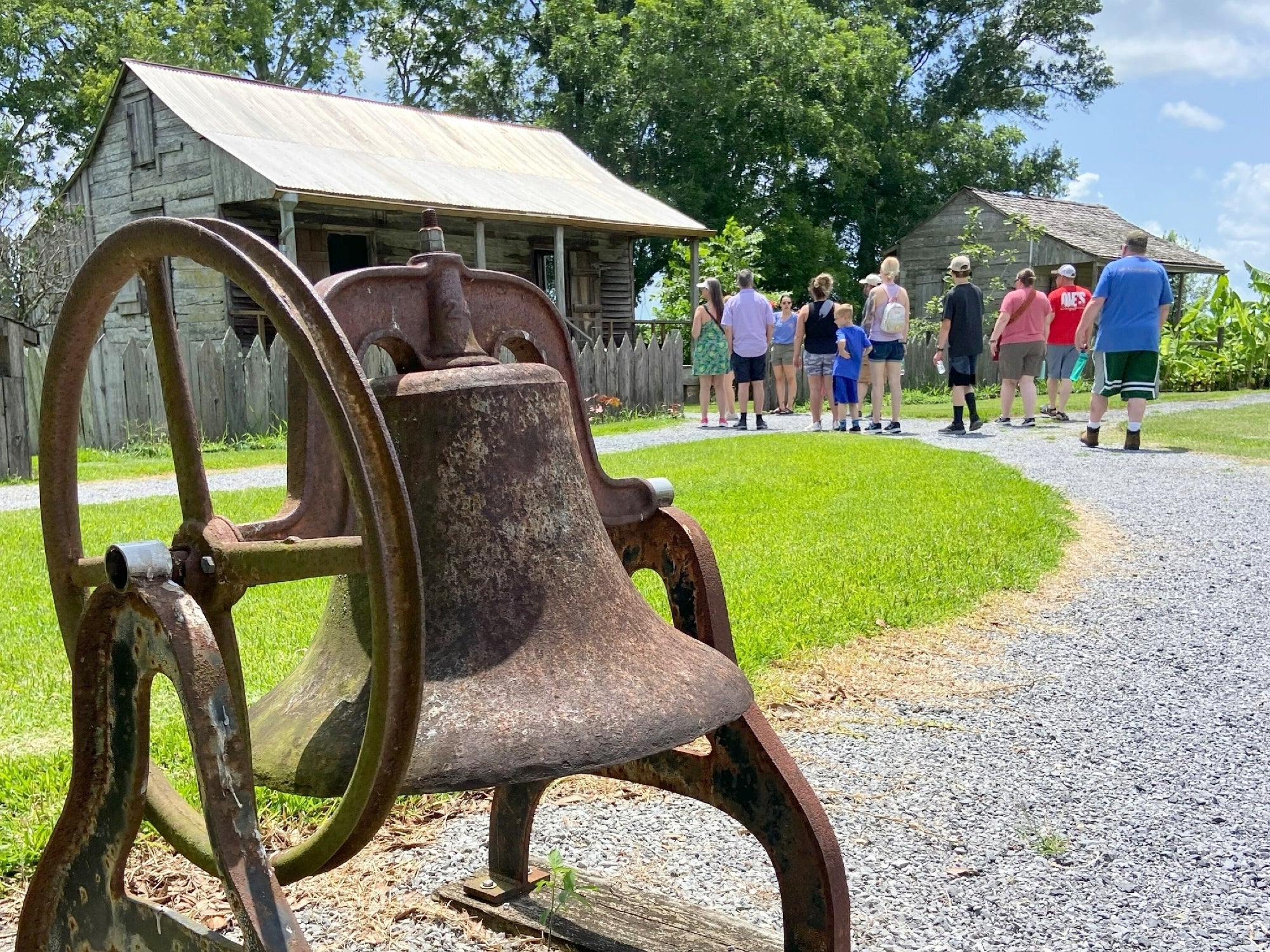 A group of people gathers near two old wooden buildings as an old, rusted bell sits in the foreground on a gravel path.
