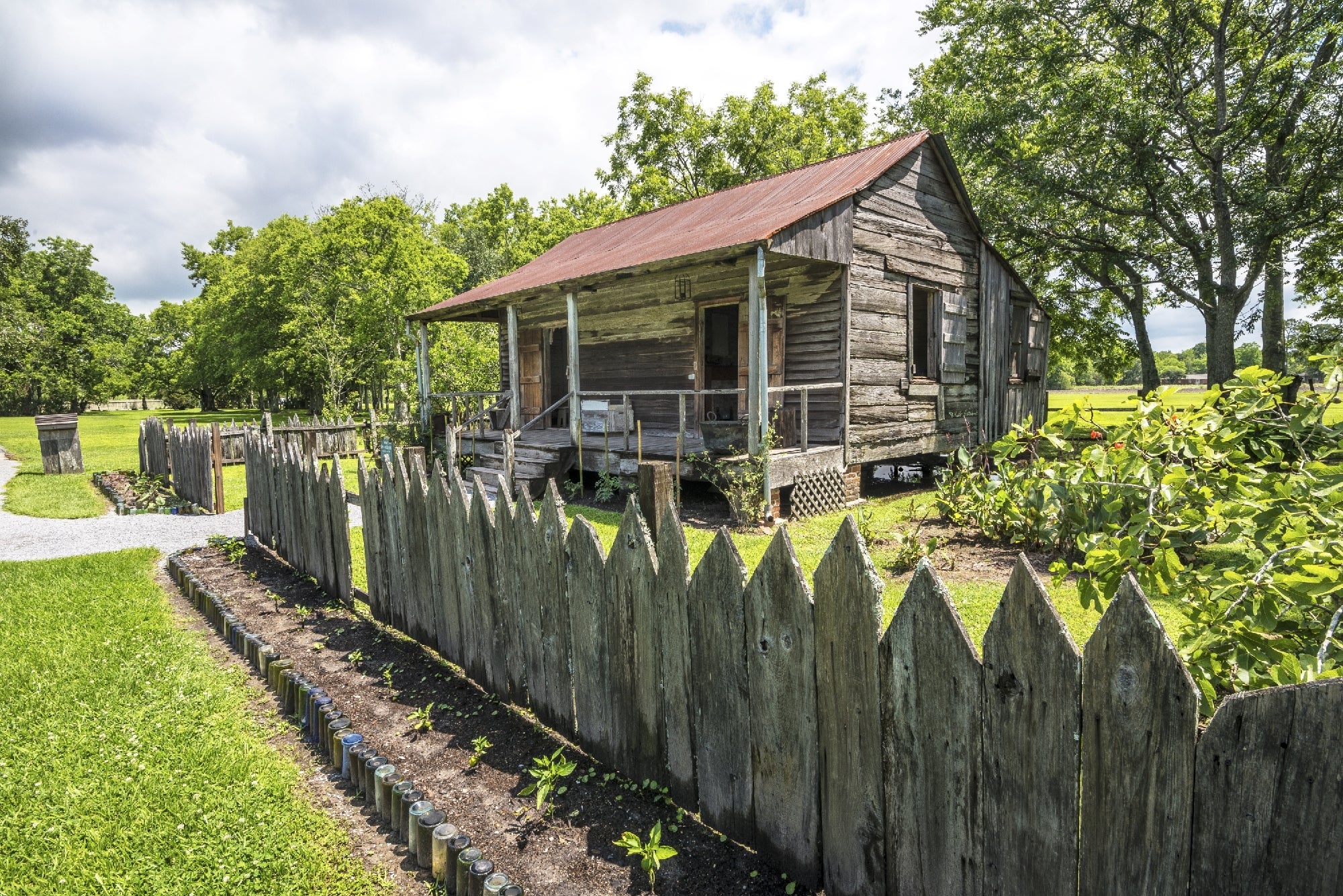 A weathered wooden shack with a rusted metal roof stands behind a jagged wooden fence, surrounded by greenery and a small garden.