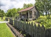 A weathered wooden shack with a rusted metal roof stands behind a jagged wooden fence, surrounded by greenery and a small garden.