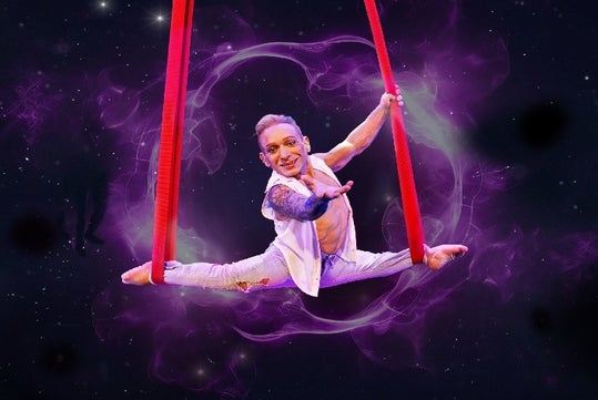Alan Silva doing the splits with one of his arms out with each of his feet in red aerial silks at Le Grand Cirque Presents Adrenaline.